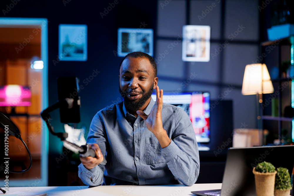 Poster enthusiastic bipoc internet show host in dark room hitting the record button on cellphone attached t