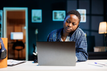 Dedicated female freelancer working from home, having business phone calls while using digital laptop. African american college student utilizes modern technology for online communication.