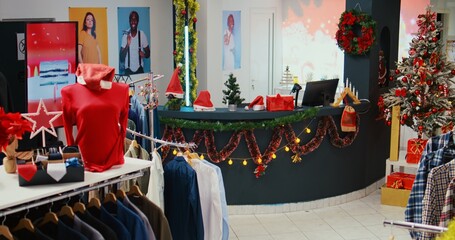 Jib down shot of festive fashion boutique filled with beautifully decorated xmas tree, garlands and merry Christmas digital screens. Empty shopping mall store with clothes on racks