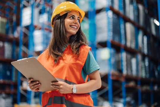 Young female warehouse auditor checks supplies with hard hat and clipboard