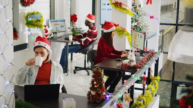 Zoom in portrait shot of happy asian employee working on christmas season in decorated office. Smiling worker solving tasks at desk in diverse xmas ornate workplace next to BIPOC colleagues