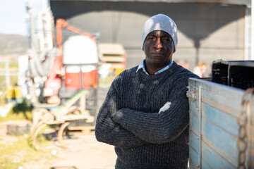 Confident adult african american farmer wearing warm knitted sweater, beanie and work gloves standing on farm on sunny spring day - Powered by Adobe