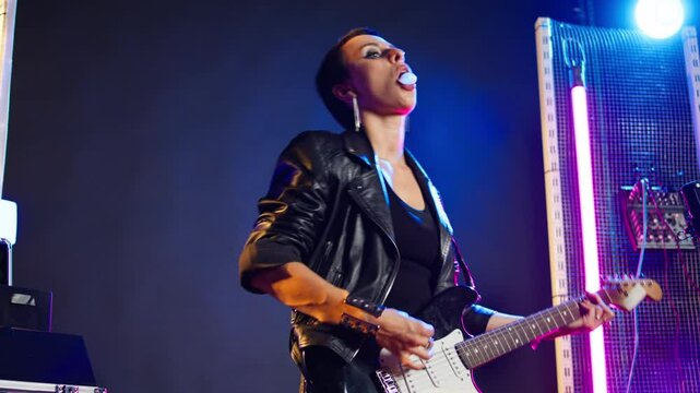 Rebellious reckless woman playing guitar and making bubbles from gum, chewing gum during her concert at the nightclub. Edgy confident rocker girl with a leather jacket. Camera A.