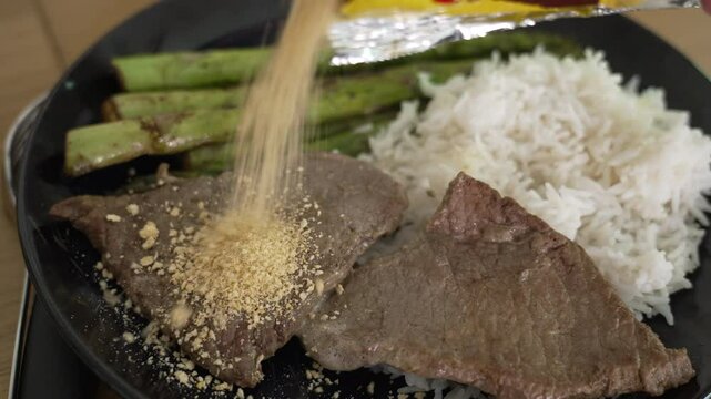 Steak garnished with farofa, a typical Brazilian ingredient, served with grilled asparagus and white rice on a black plate, highlighting a flavorful and authentic meal ideal for food photography