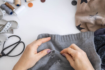 A woman uses her hands to sew up a hole in a gray knitted garment with a needle and thread. 