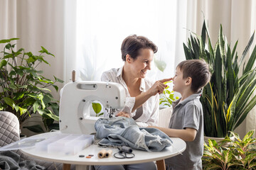 Mom and son are repairing old torn jeans using a sewing machine. A woman teaches a boy to sew