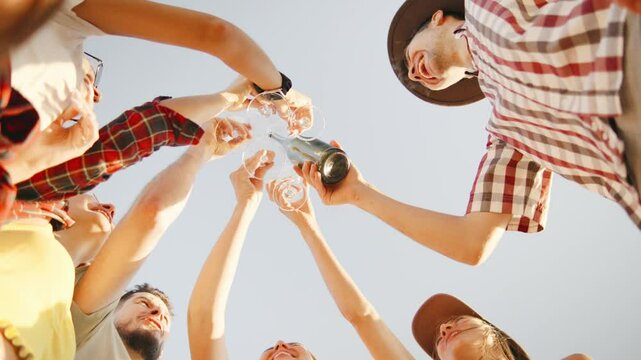 Shot from below, friends joyfully raise their glasses high into the air, make a toast and clink glasses, embodying the spirit of celebration and togetherness under a shining sunny sky.