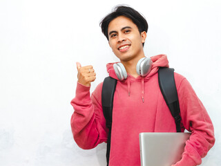 Portrait of a happy Asian male college student wearing a pink hoodie, holding a laptop, and...
