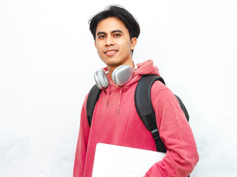 Portrait of a happy Asian male college student wearing a pink hoodie, holding a laptop, and smiling confidently while posing on an isolated white background.