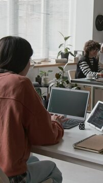 Vertical timelapse shot of busy working day of female programmer at IT startup - sitting at desk, developing computer code on laptop, testing on digital tablet, making notes, talking to colleagues