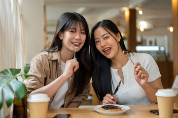 Two charming and happy Asian women are in a coffee shop, enjoying dessert and smiling at the camera.