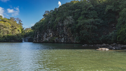 Ocean Bay. There is green tropical vegetation on the steep rocky shores. A waterfall is visible in the distance. Streams flow into the sea. Blue sky, clouds. Mauritius.Grand River South East waterfall