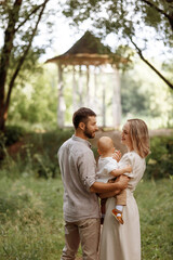 Fototapeta premium A family of three, a man and a woman with a child in their arms, stand in a park in the summer. The concept of a happy family with a small child, care and love.