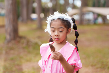 Asian girl observing a small object in hand while standing in a national park, wearing a pink dress and white headband. Child deeply focused on exploring nature and enjoying her surroundings.