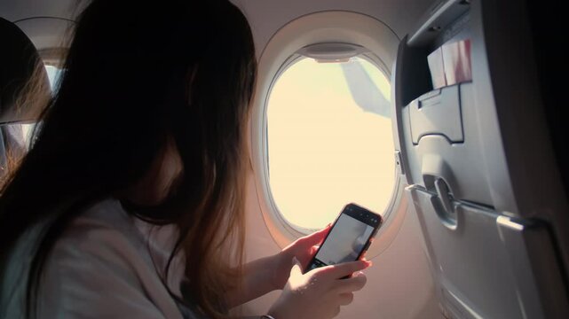 A young woman sits in an airplane and looks through the porthole and takes a photo on the phone