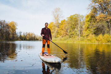 A man enjoys paddleboarding on a serene lake surrounded by the breathtaking autumn landscape,...