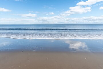 Calm blue waters meet the sandy shore on a bright summer day. The reflection of the clouds in the water creates a beautiful, peaceful scene.