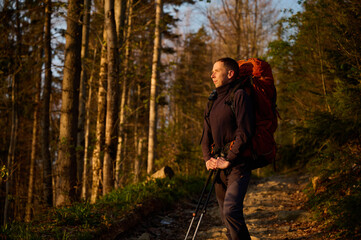 Fototapeta premium Fully equipped hiker enjoying sunrise early in the morning in the mountains. Hiker man with backpack hiking in spring forest. Hiking concept. A young traveler with a backpack walk through the woods.