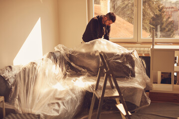 Man covering home couch with plastic sheets for protection against dust during renovation.