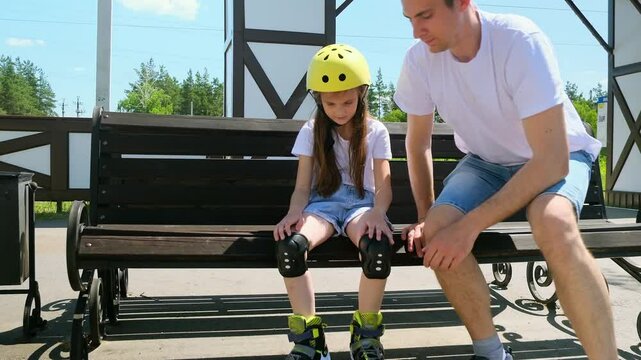 Caring father get ready the bike and roller skate safety gear, helping little adorable daughter wearing knee pad before playing friends at outdoor grass field park. Active activity, putting protective