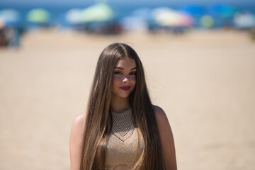 portrait of beautiful young blonde woman with long hair looks at camera happy and laughing. The girl is on the beach. In the background the blue sea and umbrellas. Concept gestures and expressions.