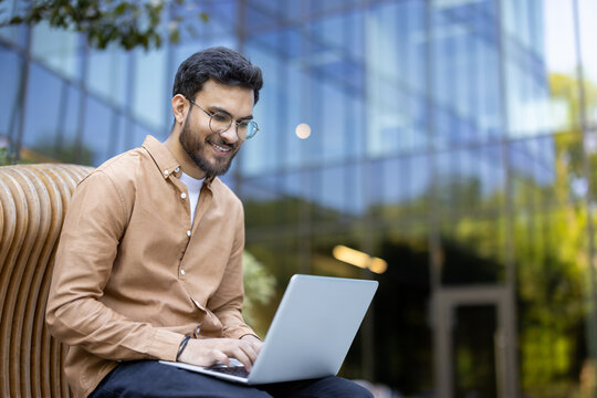 Young man smiling while using laptop outdoors in modern office environment demonstrating work flexibility and remote productivity. Business attire suggests professionalism and confidence