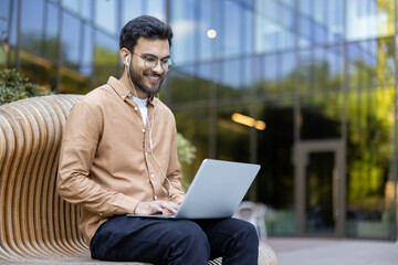 Smiling man with glasses and headphones working on laptop outdoors. Seated on stylish bench in front of modern glass building. Relaxed and professional atmosphere suggesting work-life balance