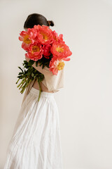 Fototapeta premium Woman in white skirt and blouse embracing large bouquet of bright red peonies. Minimalist style, elegance