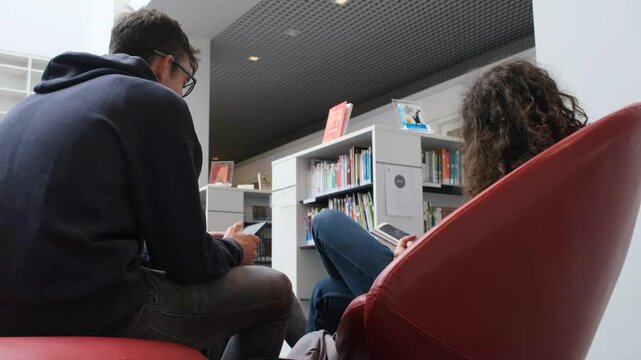 Back view of young men relaxing on library seats, consulting their smartphones or reading a comic book.