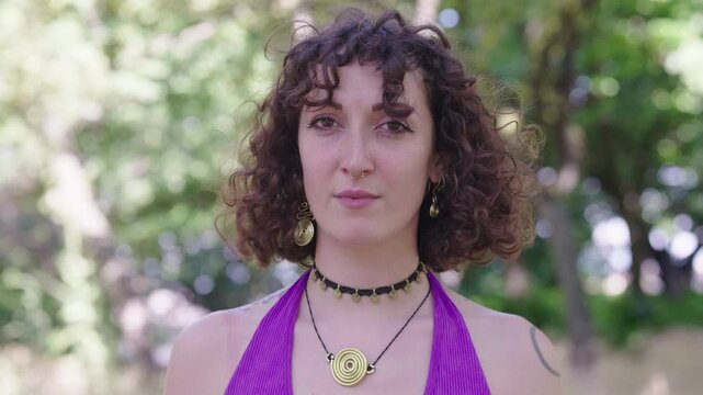 A curly-haired brunette woman with a purple shirt looks directly at the camera with a strong sense of self-awareness and confidence, capturing a moment of introspection and clarity.