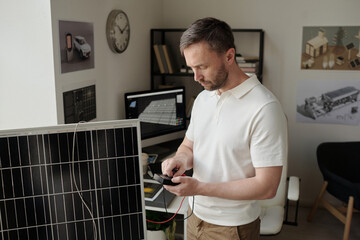 Young male engineer switching voltmeter with cable to check power while standing by solar panel