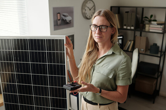 Young confident woman with ammeter standing by solar panel and checking power of alternative source of energy