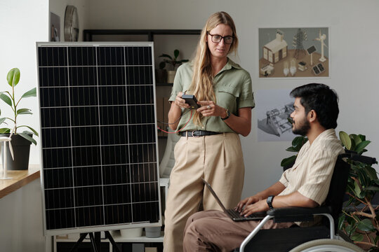 Confident female engineer with ammeter talking to male colleague in wheelchair typing on laptop keyboard