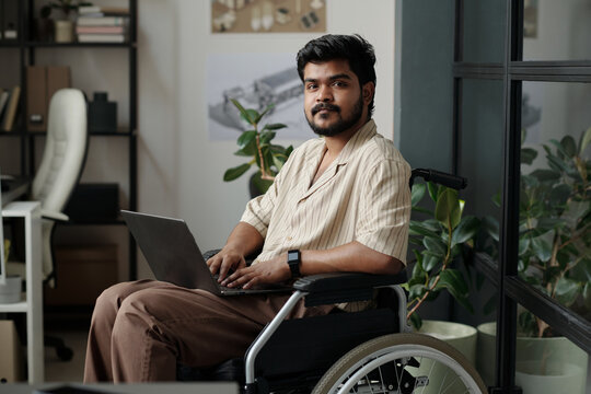 Young confident male office worker with disability sitting in wheelchair and networking while looking at camera