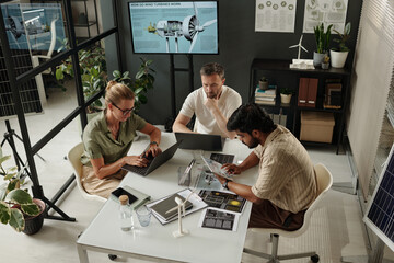 High angle of intercultural engineers sitting by desk and using mobile gadgets while working over new project