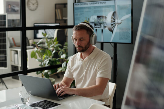 Serious male engineer in headphones typing on laptop keyboard and looking through online data on screen