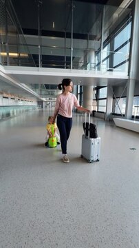 Woman pulls ride-on suitcase with her toddler son sitting on and rolling trolley case, walking through spacious, empty corridor in arrivals area of large terminal of modern international airport.