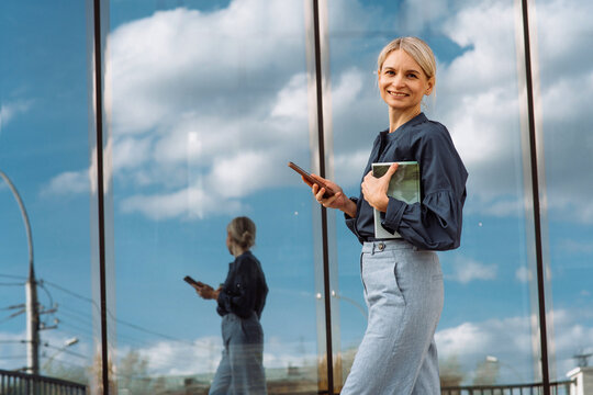 Smiling businesswoman holding smart phone and diary walking in front of glass wall