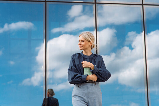 Smiling blond businesswoman with diary standing in front of glass wall