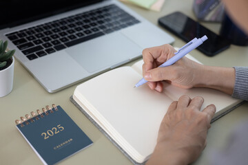 Woman is writing a plan in a notebook, year 2025 calendar on office table and work and event planning