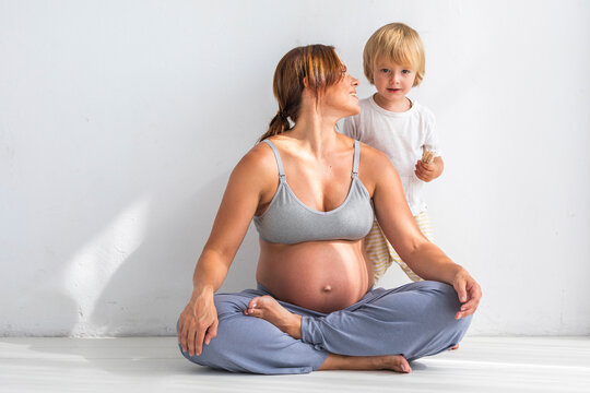 Pregnant woman sitting with cross-legged looking at son standing near white wall