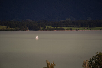 A lone sailboat floats on a calm lake, surrounded by dark forested hills and green fields in the...