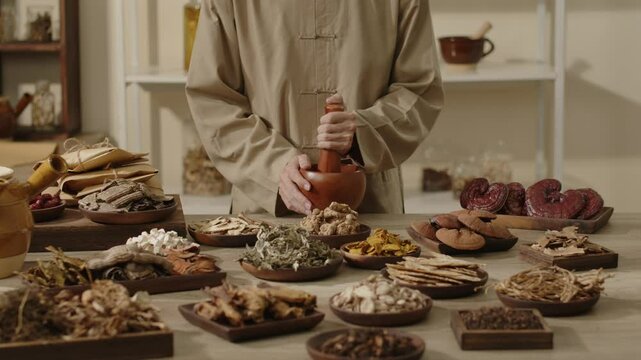 A doctor in brown traditional using a wood mortar and pestle to attire grinds dried herbs, surrounding are other herbal ingredients in a traditional laboratory setting for traditional medicine videos