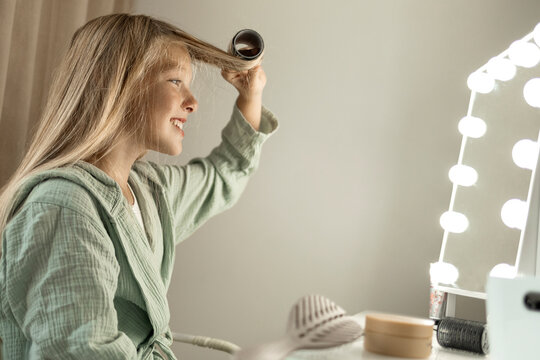 Happy blond girl curling hair in front of mirror