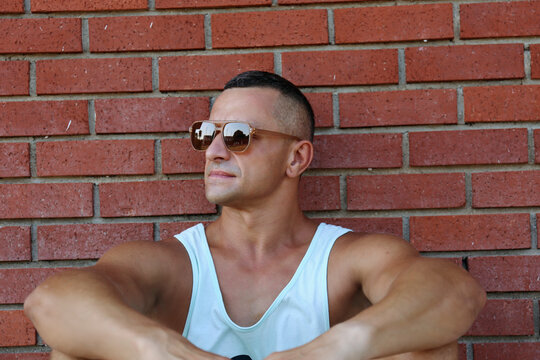 Man in sunglasses and undershirt sitting near brick wall