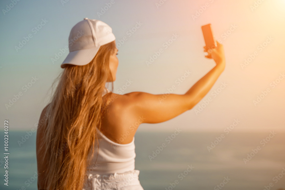 Wall mural Selfie woman sea. The picture depicts a woman in a cap and tank top, taking a selfie shot with her mobile phone, showcasing her happy and carefree vacation mood against the beautiful sea background