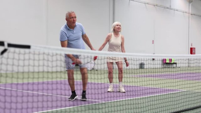 Sporty couple of senior woman and man playing doubles pickleball on indoor court, ready to hit ball. Sport and active lifestyle concept