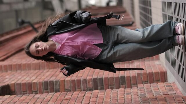 young woman leans playfully against a wall, with her leather jacket and pink shirt adding a stylish touch to her casual look. Her carefree pose reflects a fun and spontaneous attitude toward life.