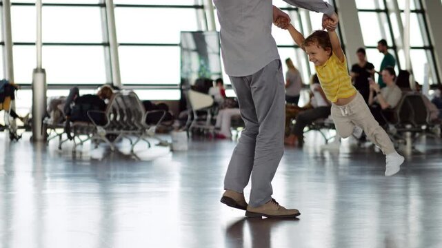 Holding his little son by hands, father spins him around, while boy laughs and kicks his legs in air. They are joyfully passing time while waiting for their flight in large airport