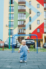 A toddler boy in denim overalls runs and plays on the playground. Active time of the child outside. A child runs on the street in the summer.